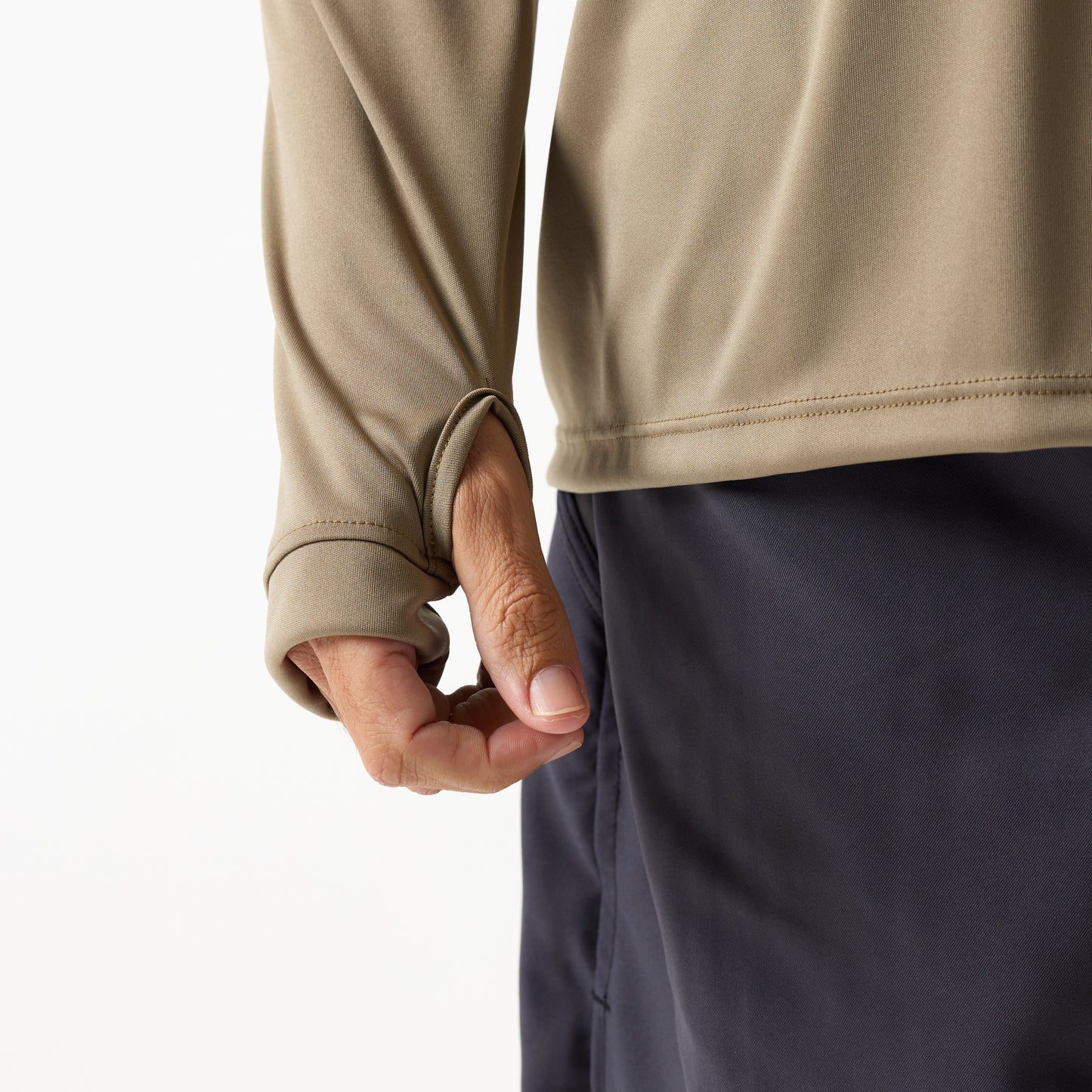 A person wears the DFND SPEED TECH HOODED 1/4 ZIP in beige, featuring a thumb hole, with dark pants and their hand partially visible through the sleeve.