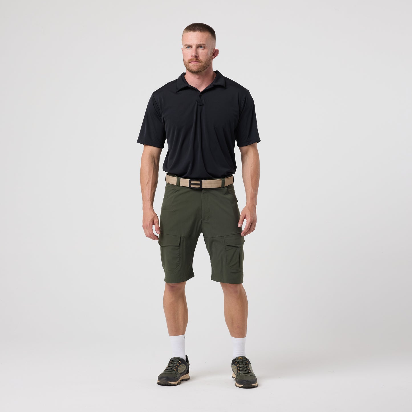 A man stands against a plain white background wearing a DFND Utility Polo with moisture-wicking technology, green cargo shorts, a beige belt, white socks, and athletic shoes.