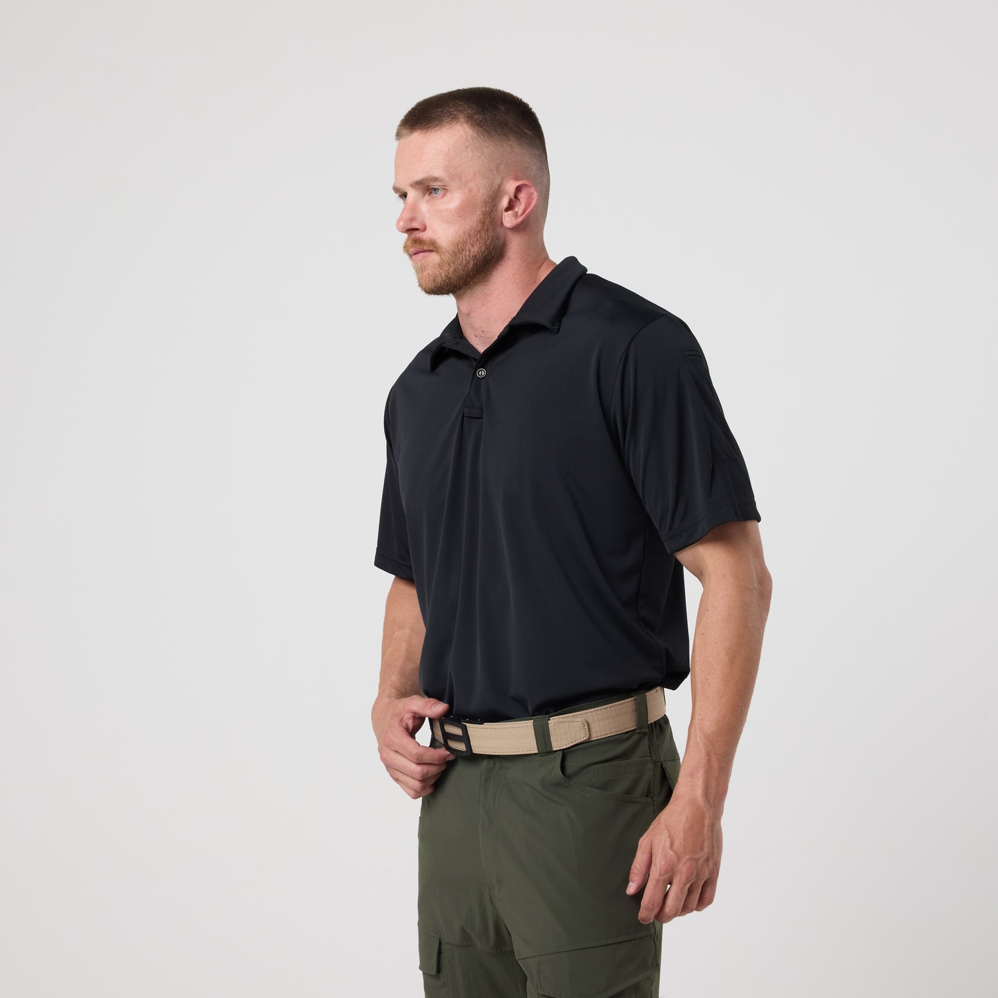 A man with short hair and a beard wears the DFND Utility Polo, featuring moisture wicking and SPF 50+ protection, in black, paired with olive green pants, standing against a plain white background.