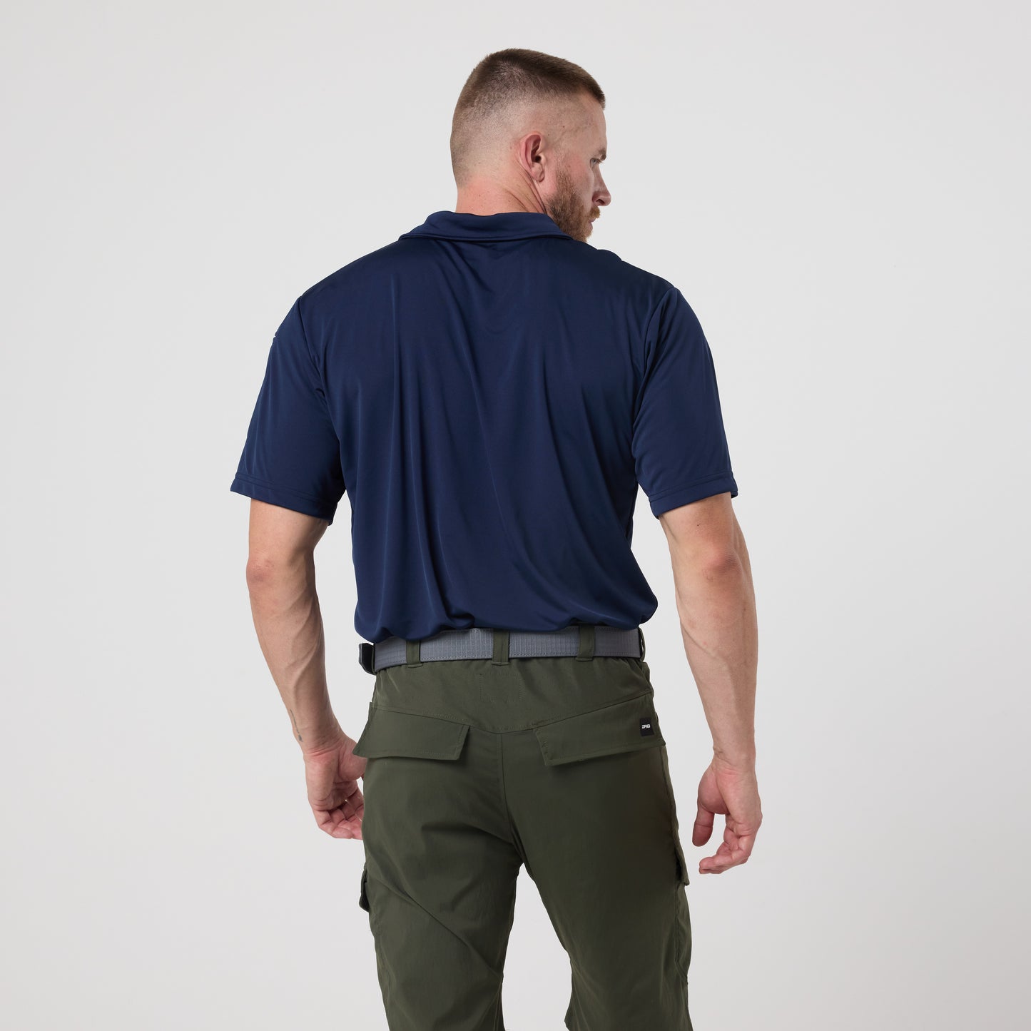 A man stands with his back to the camera, wearing the DFND Utility Polo featuring moisture-wicking technology and olive cargo pants, set against a plain white background.