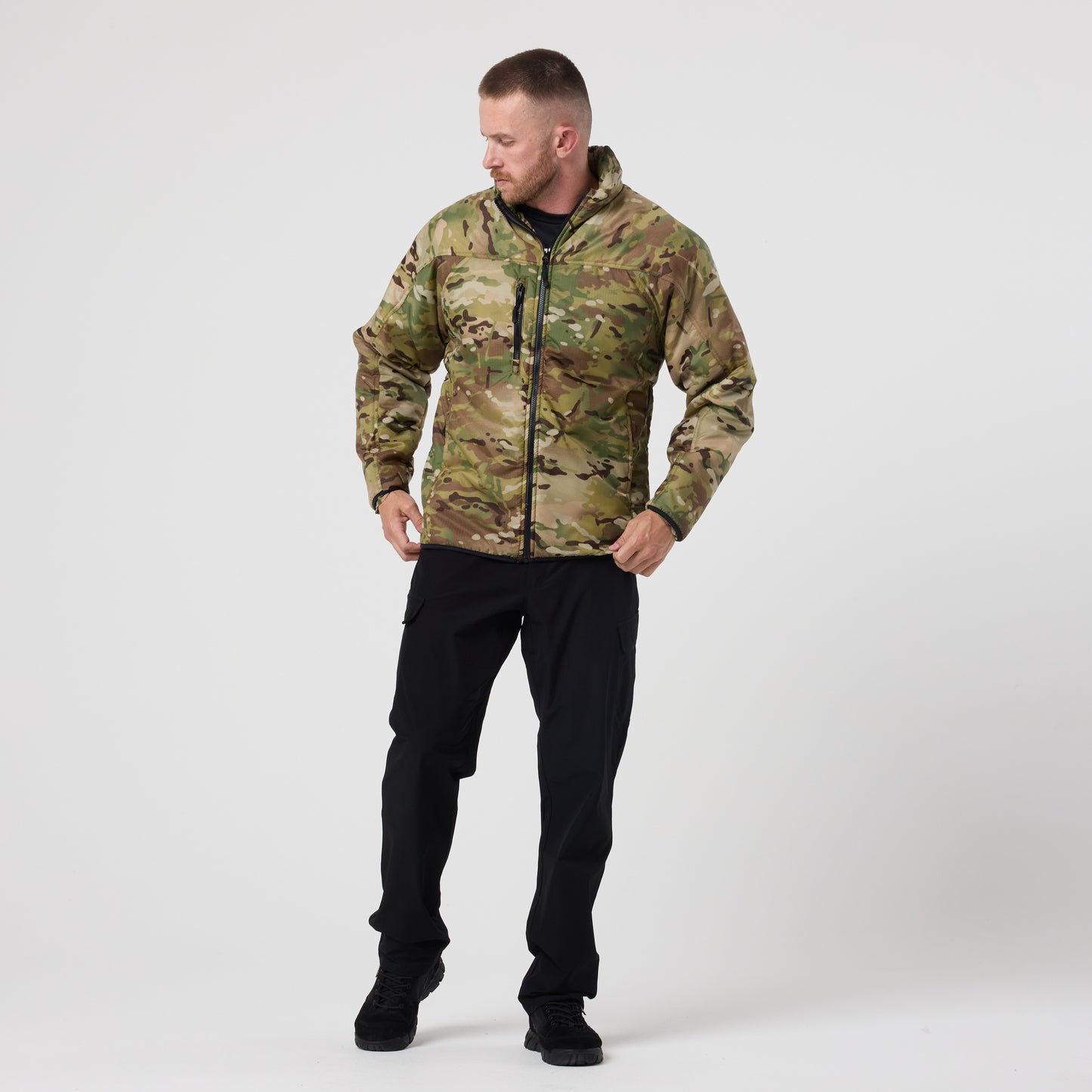 A man stands against a plain white background wearing the DFND Warrior Insulated Jacket with a DWR shell and black pants, looking down and to his left.