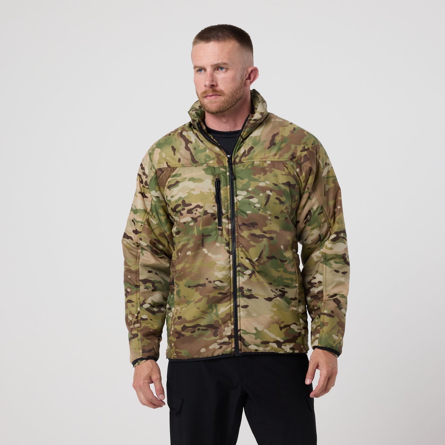 A man wearing a DFND Warrior Insulated Jacket with a camouflage pattern and black pants stands against a plain light gray background.