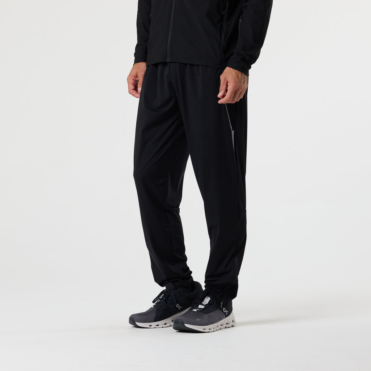 A person wearing DFND FLASH TECH PANTS featuring 4-Way Stretch, along with a black jacket and black running shoes, stands against a plain white background.