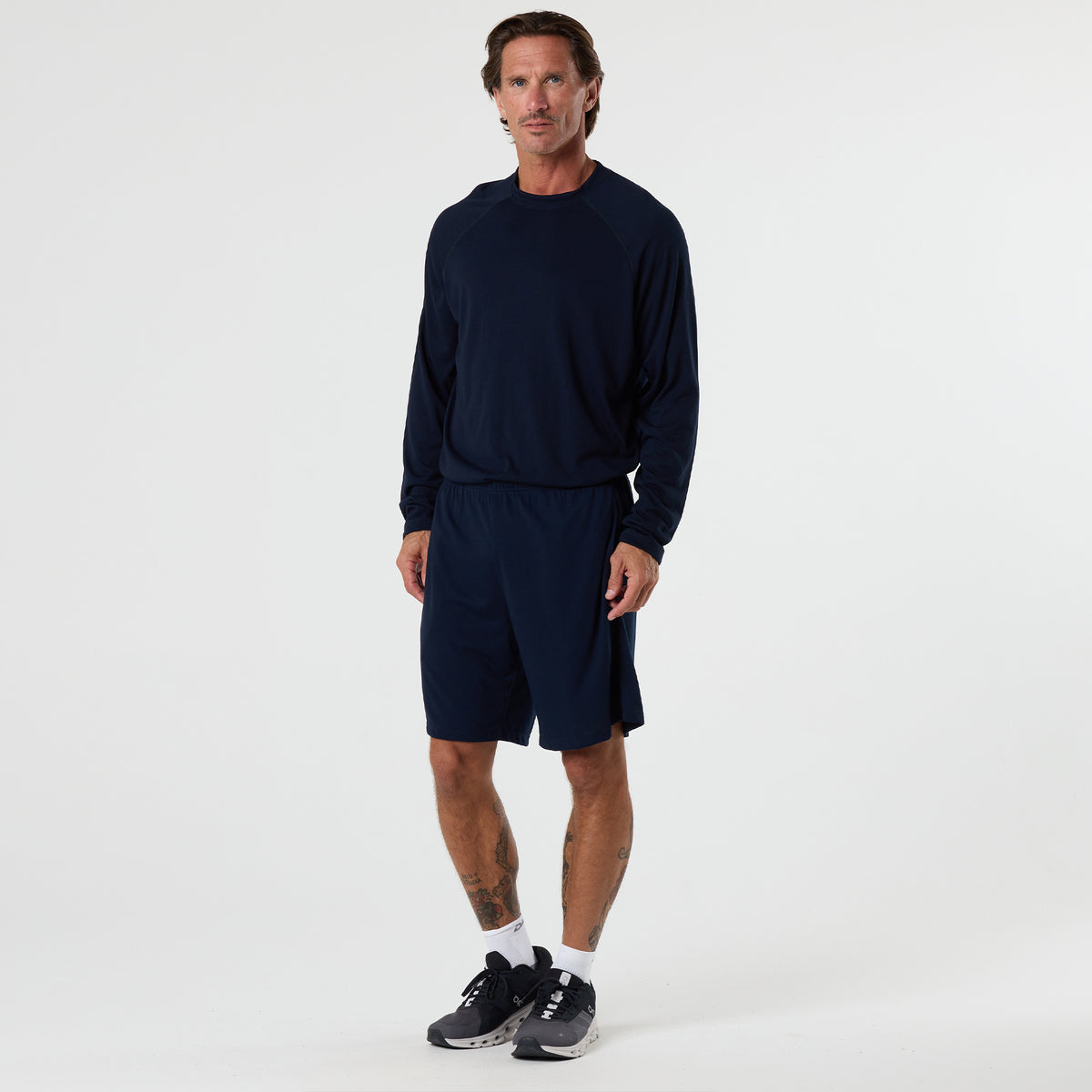 A man stands against a plain white background wearing the DFND FR Performance Long Sleeve Shirt with raglan sleeves in navy, paired with navy shorts, white socks, and black athletic shoes.