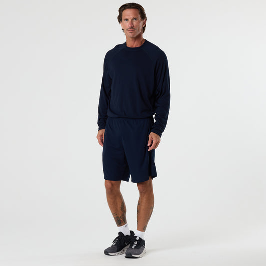 A man stands against a plain white background wearing the DFND FR Performance Long Sleeve Shirt with raglan sleeves in navy, paired with navy shorts, white socks, and black athletic shoes.