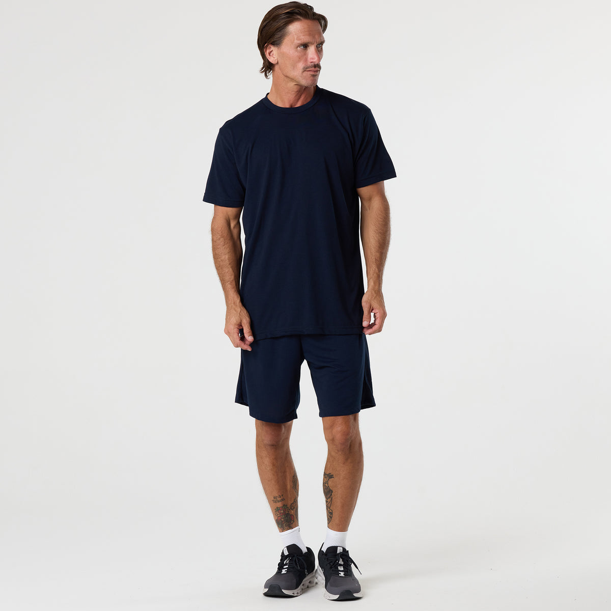 A man stands in a studio wearing the DFND FR PERFORMANCE SS SHIRT - Traditional Sleeve in navy, designed as a lightweight base layer, paired with matching shorts and black sneakers. He looks to the side with arms relaxed.