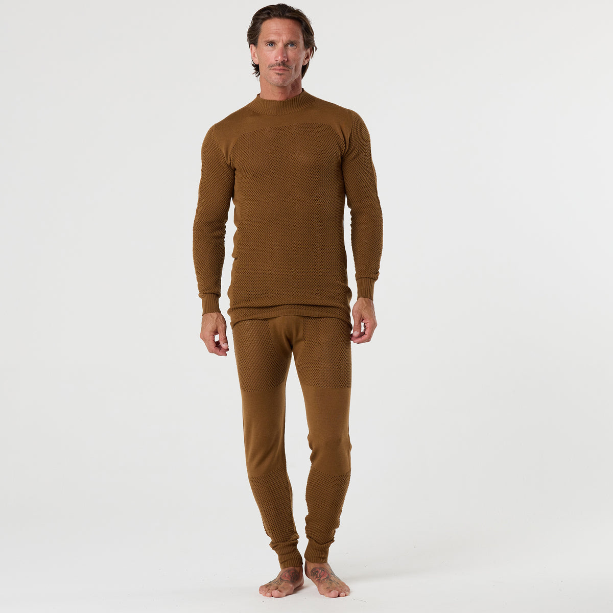 A man stands barefoot on a white background, facing the camera, wearing the DFND TAHOE WOOL MESH LONG SLEEVE SHIRT and matching pants set in brown. This textured outfit is designed for cold weather performance and functions perfectly as a wool mesh base layer.