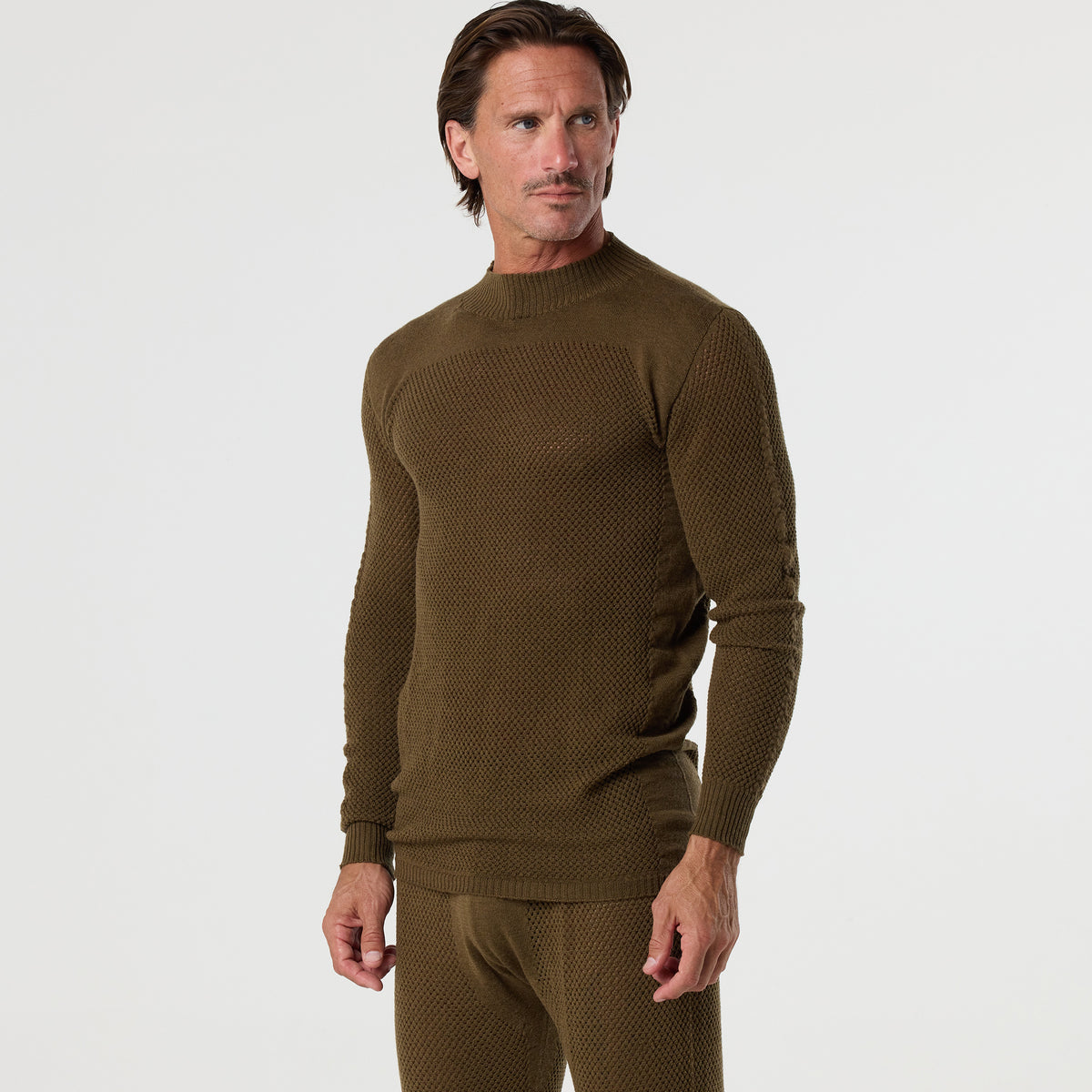 A man wears the DFND TAHOE WOOL MESH LONG SLEEVE SHIRT in olive, paired with matching pants, designed for cold weather performance; he stands against a plain white background while looking to his left.