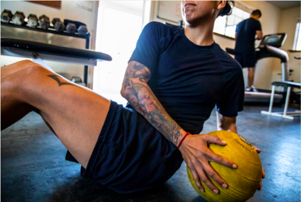 Person exercising with a yellow medicine ball in a gym, sitting on the floor with a tattooed arm visible. Another person is on a treadmill in the background.
