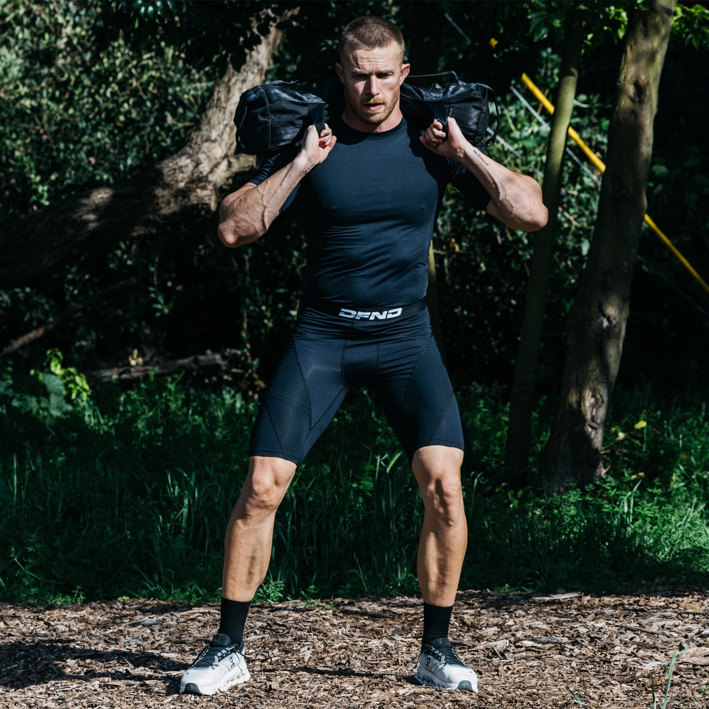 A man in DFND's Compression Short + Shirt Gym Kit stands outdoors on a dirt and wood chip surface, holding a sandbag across his shoulders as he prepares to exercise.