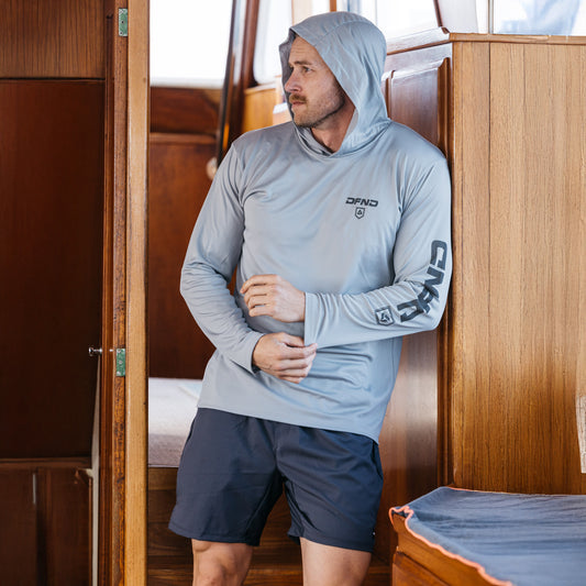 A man in a light gray DFND Speed Tech Hoodie and Aero Short Sun Protection Kit stands indoors on a wooden boat, looking to his left.