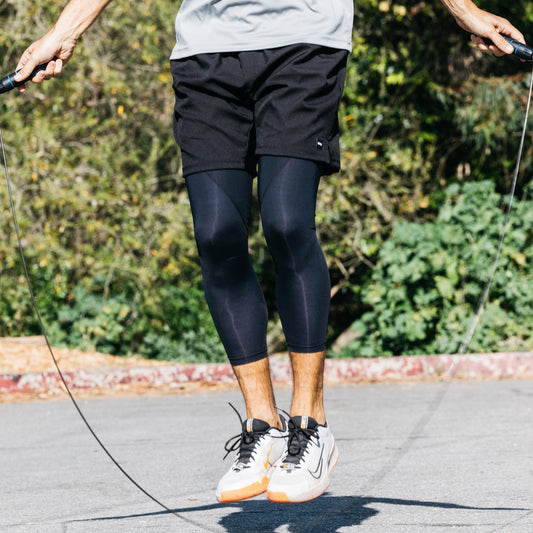 Person skipping rope outdoors, wearing a gray shirt, black DFND AERO SHORT 7", black athletic leggings, and white sneakers. Trees and shrubbery appear in the background.