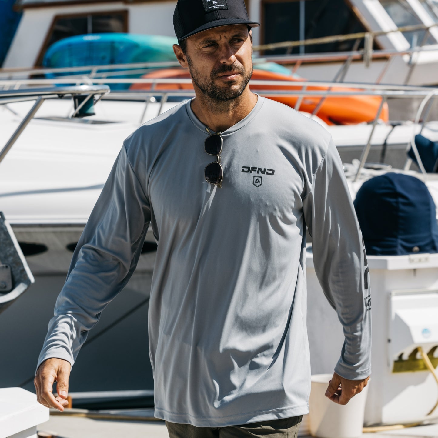A man in a gray DFND SPEED TECH LS SHIRT W/DFND LOGOS and black cap walks at a marina, sunglasses hanging from his collar.